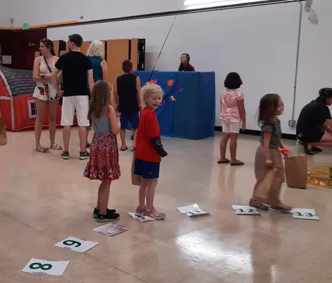 Children participate in an indoor activity while adults and other children stand around them.