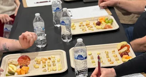 People sitting around a table with trays of cheese samples, water bottles, and clipboards.