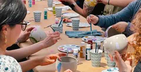 People sit around a long table painting pottery bowls and objects during an art class.