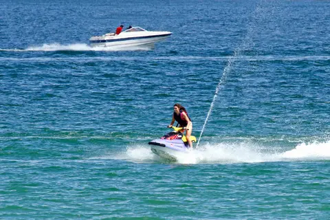 A person rides a personal watercraft on blue water with a motorboat in the background.
