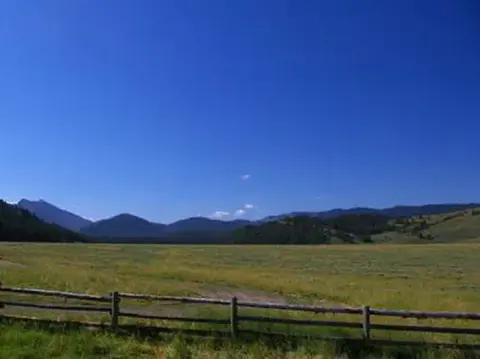 A scenic landscape showing a wide grassy field with a wooden fence in the foreground and mountains under a blue sky in the background.