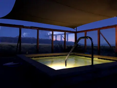 An illuminated hot tub sits inside an open-air structure at dusk with mountains visible in the distance.
