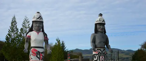 Two carved wooden figures stand outdoors against a cloudy sky and distant hills.