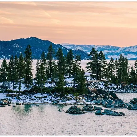 A scenic view of a snowy forested shoreline alongside a body of water at sunset.