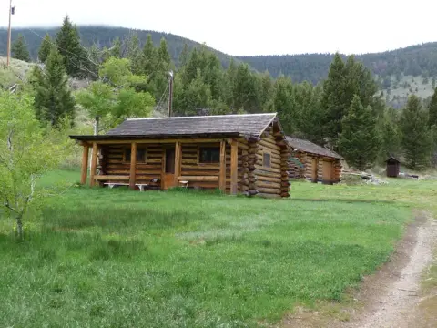 A log cabin sits in a grassy clearing with a wooded hill in the background.