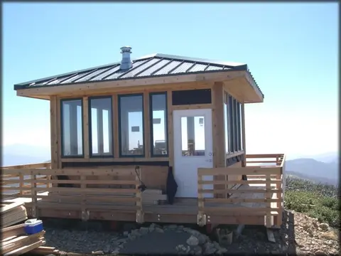 A wooden lookout cabin sits atop a mountain under a clear blue sky.