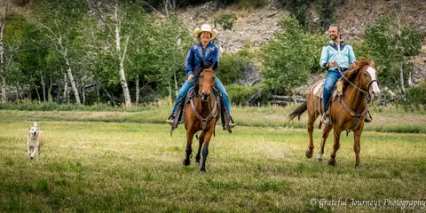 Two people ride horses across a grassy field with a dog running alongside.