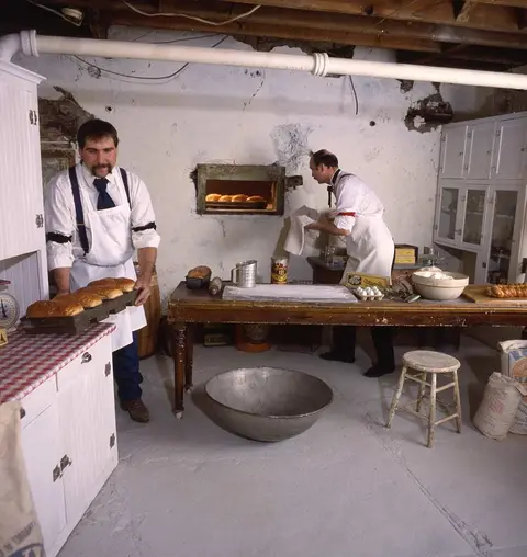 Two bakers in period attire work inside a rustic bakery.