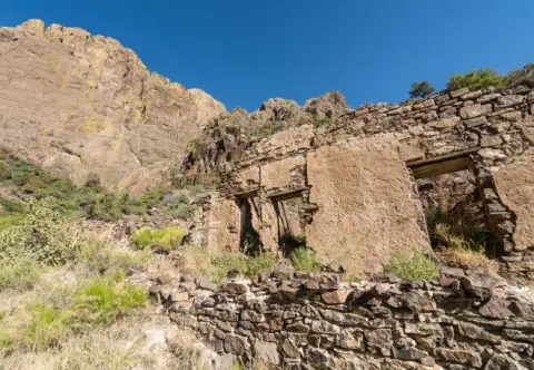 Stone ruins sit below a rugged mountain slope under a blue sky.