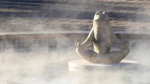 A frog statue in a yoga pose sits on a platform in a steaming pool.