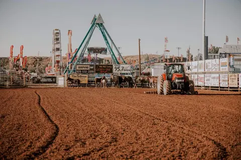 A tractor is in the dirt arena of a fair with carnival rides and horses in the background.