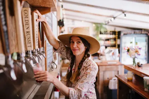 A woman in a hat pours a drink from a tap handle inside a rustic shop.