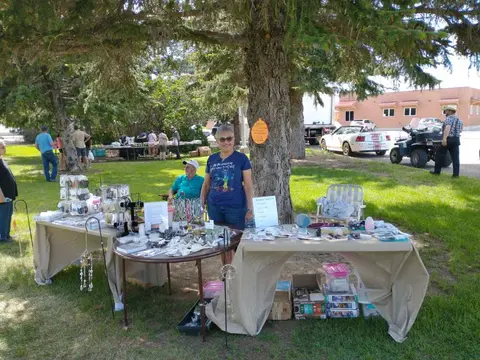 A woman stands behind tables displaying crafts and goods at an outdoor market.