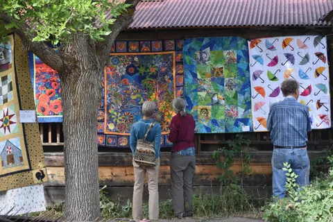 People view colorful quilts hanging on a wooden building outdoors.