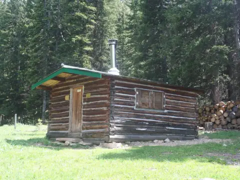 A rustic log cabin stands in a clearing surrounded by a dense evergreen forest.
