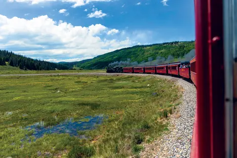 A red passenger train travels along a curve through a green landscape with mountains and a cloudy blue sky.