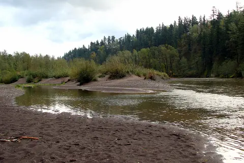 A river flows past sandy banks and forested hills under a cloudy sky.
