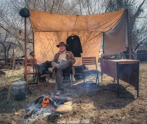 A man sits in front of a tent and campfire.