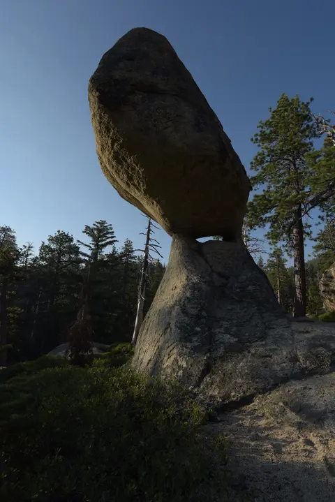 A large balancing rock formation stands in a forest under a clear sky.