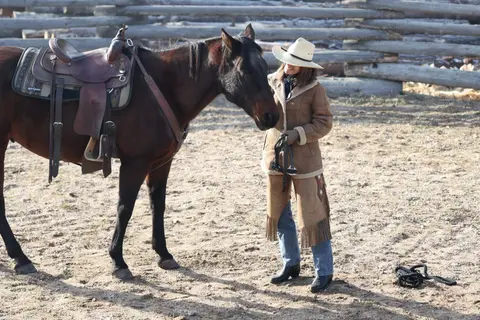 A woman in cowboy attire stands next to a saddled horse.