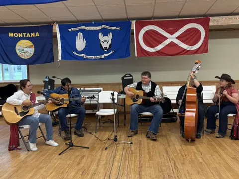 A group of five musicians play acoustic instruments indoors beneath flags.
