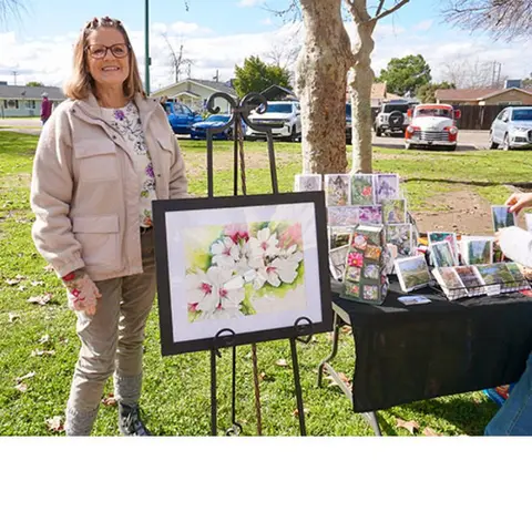 An artist stands beside her framed painting at an outdoor market.