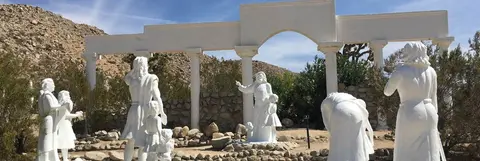 White statues of figures are arranged outdoors in front of a white colonnade structure in a desert setting.