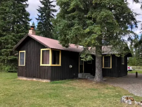 Exterior view of a dark wood cabin with yellow-trimmed windows and a reddish roof surrounded by trees.