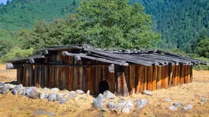 A rustic wooden plank building stands before a forested mountain.