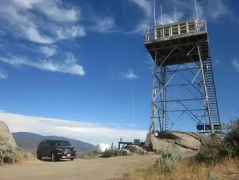 A tall metal lookout tower stands next to a parked car on a dirt road.