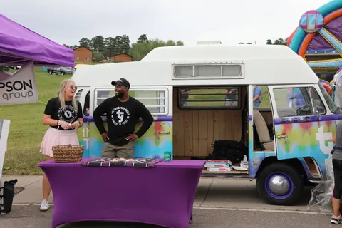 Two people stand next to a customized retro van and a table outdoors.