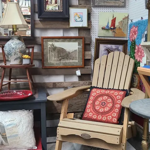 An indoor display features a wooden chair with a patterned pillow, surrounded by framed artwork, a lamp on a small table, and other decorative items.