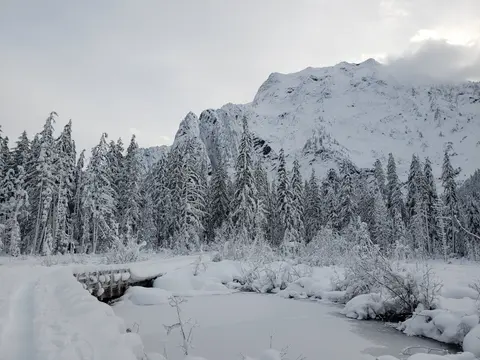 A snowy landscape features a frozen pond, a small wooden bridge, and snow-covered trees beneath a massive mountain.