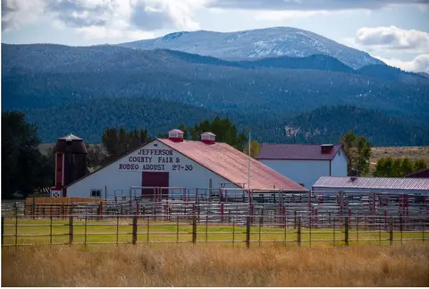 A large fair and rodeo barn with metal pens is set against a mountain backdrop.