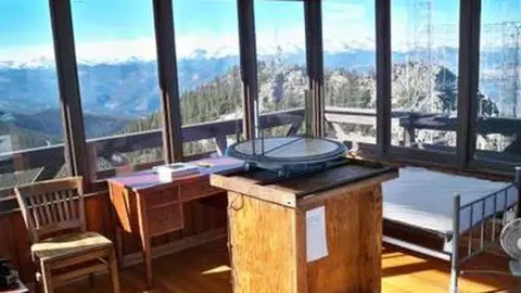 Interior view of a fire lookout tower with windows showing mountain views, a desk, and a finder instrument.