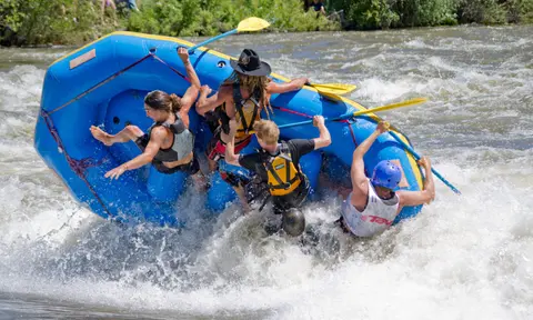 A blue raft is tipping over in whitewater rapids with people inside.