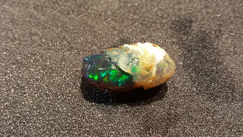 A raw opal gemstone with vibrant green and blue fire sits on a textured dark surface.