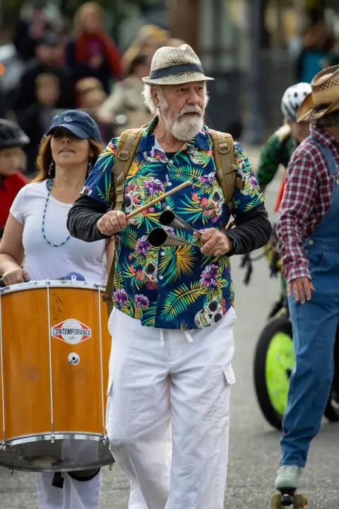 A man wearing a patterned shirt and hat plays percussion instruments while walking in a street event.