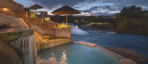 A hot spring pool with stone terraced walls and umbrellas beside a river at dusk.