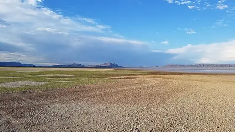 A wide landscape showing a dry flat plain with sparse green vegetation and distant hills under a cloudy blue sky.