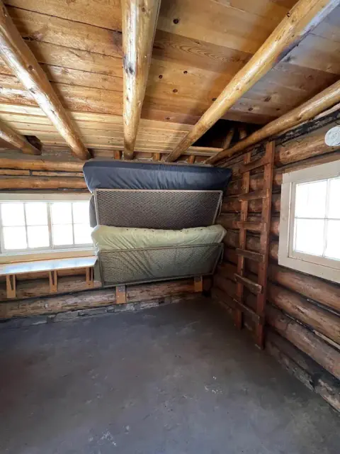 An interior view of a rustic log cabin with bunk beds and a wooden ladder.