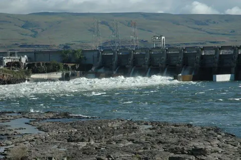 A large dam releases water into a rushing river beneath rolling hills.