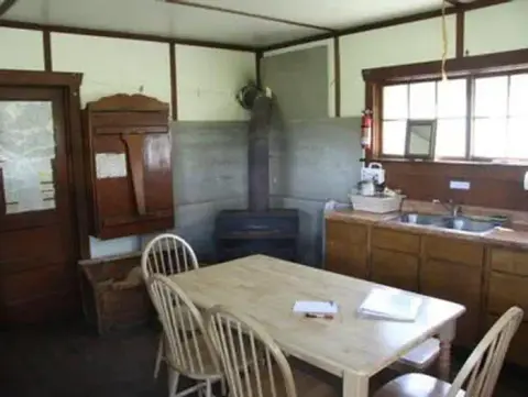 A rustic interior showing a dining table, chairs, wood stove, and kitchen counter.