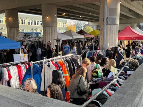 A busy outdoor flea market or clothing fair set up under a highway overpass.