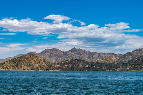 A scenic view of a body of water with hills and mountains under a blue sky with clouds.