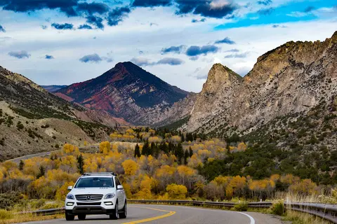 A white SUV driving on a mountain road through a valley with autumn trees.