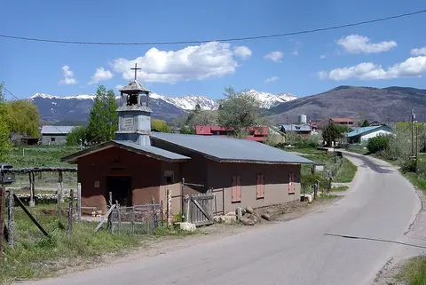 An old adobe church stands beside a road with snow-capped mountains in the background.