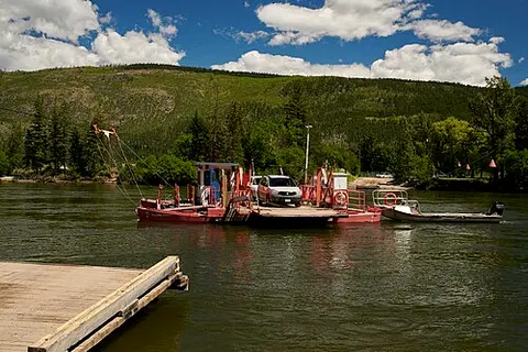 A small ferry boat carrying cars on a river with green hills in the background.