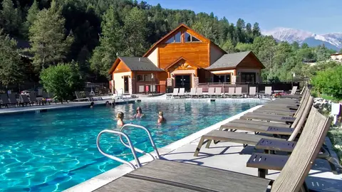 People swimming in an outdoor pool with lounge chairs and a wooden building in the background.