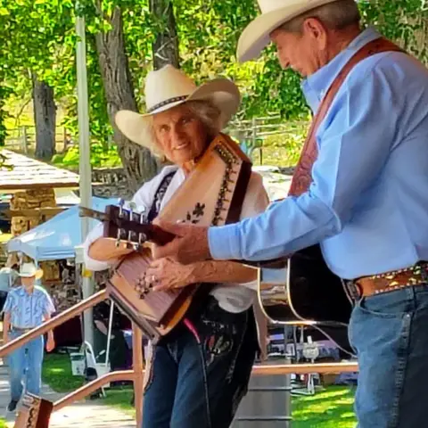 Two people wearing cowboy hats play stringed instruments outdoors.